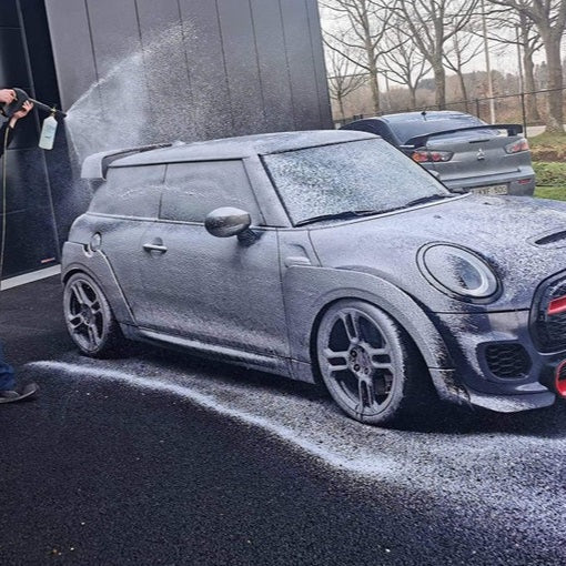 Person washing a Mini Cooper with a hose on a driveway.